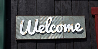 Photo shows a handwritten sign with Welcome on a dark gray plank wall