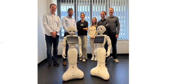 The image shows a group of six people in a bright office standing side by side behind two identical, white, humanoid robots. The two robots in the foreground feature a rounded base that widens at the bottom, slender bodies, articulated arms, and heads with large, pink-glowing eyes, along with a landscape-oriented tablet mounted on each of their chests. Behind them, standing from left to right, are four men and two women: a man in a light blue shirt and dark jeans, a man wearing glasses in a light blue shirt and dark pants, a woman with crossed arms in a black top and wide, light grey pants, a woman in a light beige jacket and white pants, a man in a dark shirt, and a man in a grey sweater. The group is standing in front of a large window with partially closed vertical blinds and a radiator underneath. The room has white walls and a dark, slightly speckled floor, with some cables visible on the ground in the right background.