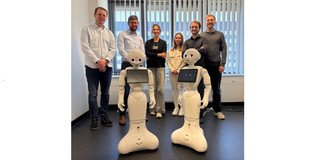 Das Bild zeigt eine Gruppe von sechs Personen in einem hellen Büro, die nebeneinander hinter zwei identischen, weißen, humanoiden Robotern stehen. Die beiden Roboter im Vordergrund haben eine abgerundete Basis, die nach unten hin breiter wird, schmale Körper, Gelenkarme und Köpfe mit großen, rosafarben leuchtenden Augen sowie jeweils ein querformatiges Tablet, das auf ihrer Brust montiert ist. Hinter ihnen reihen sich von links nach rechts vier Männer und zwei Frauen auf: ein Mann in hellblauem Hemd und dunkler Jeans, ein Brillenträger in hellblauem Hemd und dunkler Hose, eine Frau mit verschränkten Armen in schwarzem Oberteil und hellgrauer, weiter Hose, eine Frau in heller, beiger Jacke und weißer Hose, ein Mann in dunklem Hemd sowie ein Mann in grauem Pullover. Die Gruppe steht vor einer großen Fensterfront mit teilweise geschlossenen vertikalen Lamellenjalousien, unter der sich ein Heizkörper befindet. Der Raum hat weiße Wände und einen dunklen, leicht gesprenkelten Fußboden, auf dem auf der rechten Seite im Hintergrund einige Kabel liegen.