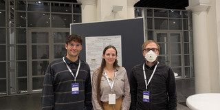 The image displays a group photo of three individuals standing side-by-side indoors, likely at an academic conference or symposium. They are positioned in front of a dark grey display board holding a research poster with the partially visible title "Fighting apps with apps: Analyzing tools for digital minimalism." The person on the left is a man with short, dark curly hair and glasses, wearing a dark grey sweater with thin horizontal stripes. In the center stands a woman with long, straight brown hair and glasses, dressed in a light-colored striped blouse and tan trousers. On the right stands a man with reddish-blond hair and glasses, wearing a dark button-down shirt and a white FFP2 face mask covering his nose and mouth. All three individuals are wearing blue lanyards with conference badges around their necks and are looking directly at the camera. The background reveals the architecture of a spacious hall featuring high white columns and a large wall of glass-paneled doors or windows, looking out into a darker area. A small portion of a white high-top table is visible in the bottom right corner of the frame.
