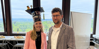 The image shows a woman and a man standing next to each other in a brightly lit room, smiling directly at the camera. The woman on the left has long blonde hair and is wearing a light pink pantsuit with a black top underneath, a gold necklace, and black pointed shoes. On her head, she wears a striking, custom-decorated doctoral hat. The cylindrical base of the hat is covered with several printed photos, while the square top features a small rubber duck wearing its own miniature graduation cap and a green tassel. Standing to her right is a man with short brown hair, a beard, and round glasses. He is dressed in a white button-down shirt, a gray and black patterned blazer, light beige trousers, a dark belt, and brown leather brogue shoes; he wears a watch on his left wrist. In the background, large windows offer a sweeping view of a lush green, forested landscape under a cloudy sky. To the left side of the room, there is a high table set up with platters of snacks and finger food, with a red plastic crate sitting on the floor underneath it. On the right side, a mobile whiteboard on wheels and parts of other office equipment are visible. The light-colored floor is marked in a few places with strips of yellow tape.