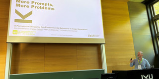 A presenter stands at a black lectern marked with a white "JKU" logo in a wood-paneled lecture hall. He is wearing a striped button-down shirt, a conference lanyard, and holds a slide clicker in his right hand. To his left, a large projection screen displays a title slide: "More Prompts, More Problems – A Persuasive Design for Pro-Environmental Behaviour in Image Generations" by Kay Hönemann, Fabian Helms, and Manuel Wiesche from TU Dortmund University. The slide footer references the International Conference on Information Systems (WI) at JKU Linz and includes the JKU Business School logo. In the foreground, a desk holds monitors, input devices, water bottles, and hand sanitizer. On the far right, a tall window looks out onto greenery.