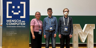 Three researchers stand side by side in front of a green wall at the Mensch und Computer 2026 conference. On the left is a woman in a pink shirt, in the center a man with glasses in a patterned shirt, and on the right a man with glasses, an FFP2 mask, and a dark shirt. All three wear conference lanyards. To their left is a blue banner reading "Transforming Interactions", and to their right is a large white illuminated letter "M".