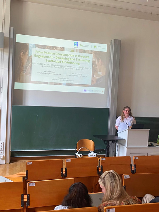 Eine Aufnahme aus der Perspektive eines Zuschauers in einem Hörsaal, die eine Frau bei einer Präsentation zeigt. Sie steht rechts im Bild an einem weißen Rednerpult, blickt konzentriert auf ihren Laptop und trägt das gleiche weiße Oberteil mit Lochstickerei wie auf dem Gruppenbild. Über ihr wird eine Präsentationsfolie groß auf eine Leinwand projiziert. Die Folie trägt den Titel „From Passive Consumption to Creative Engagement - Designing and Evaluating Scaffolded AR Authoring“ und listet darunter die Namen Dana Hofmann, Kay Hönemann und Manuel Wiesche samt E-Mail-Adressen auf. Hinter der Rednerin befindet sich eine breite grüne Tafel, davor steht ein Tisch mit einem Beamer. Im Vordergrund sind die braunen hölzerner Sitzreihen des Hörsaals zu sehen, auf denen einige Zuschauer sitzen, deren Hinterköpfe und Schultern im unscharfen Bereich des Bildes liegen.