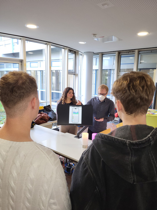 The vertical photograph captures a conversation scene in a brightly lit, modern room with large windows, likely during an exhibition or open house event. In the foreground, two young men stand with their backs to the camera, facing a white presentation table. The man on the left has short light-brown hair and is wearing a white cable-knit sweater. The man to his right has dark brown hair and wears a dark grey or black hooded jacket made of sturdy fabric.  Behind the table, two presenters face the visitors. On the left stands a young woman with long, curly brown hair, wearing a dark top and beige trousers; she is looking towards the presentation screen. Next to her stands a man with short blond hair and glasses, wearing a dark button-down shirt and a white FFP2 face mask. He holds a tablet in his hands and appears to be explaining something. Centered on the table is a black computer monitor displaying a graphic of a smartphone interface featuring various colorful bar charts and statistics. Two small, cylindrical decorative lamps sit in front of the monitor; the left one glows warm white, while the right one glows purple. Beneath the table, a tangle of cables and a blue cable reel are visible. In the background, natural light floods in through floor-to-ceiling windows, revealing the facade of a neighboring building with white and blue panels. To the left in the background, jackets and bags are piled on a windowsill or ledge.