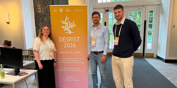 A group photo of three people standing next to a large, orange-to-pink gradient banner in a foyer. The banner reads "DESRIST 2026" in large letters and promotes the "21st International Conference on Design Science Research in Information Systems and Technology" taking place from June 8-10, 2026, in Münster. To the left of the banner stands a woman with long, light hair, wearing a white short-sleeved top with eyelet embroidery and a dark skirt. To the right of the banner stand two men: the one on the left wears glasses, a light blue button-down shirt, jeans, and a brown belt; the one on the right is the man from the first image, wearing a dark blue sweater over a white shirt and light trousers. All three are wearing conference name badges around their necks and are smiling at the camera. In the background, glass double doors lead outside to greenery, and a table with a monitor is visible on the left edge of the image.