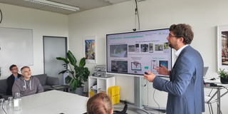 The landscape-format photo captures a presentation taking place in a bright, modern seminar or meeting room. On the right side of the image, a male presenter stands in profile, facing left towards the audience. He has short, curly brown hair, wears glasses, a blue blazer over a white shirt, and blue jeans. He is gesturing with both hands whilst holding a presentation clicker in his right hand. Behind him is a large digital flat-screen display mounted on a rolling stand. The slide being presented features the green headline "Virtuelle Gärten – Einsatzmöglichkeiten" (Virtual Gardens – Application Possibilities) and shows the logo of TU Dortmund in the top right corner. The slide content is divided into four quadrants displaying images and graphics related to Augmented Reality (AR) and Virtual Reality (VR) applications on smartphones and computer monitors.  On the left side of the image, the audience is seated at a configuration of white tables. In the immediate foreground, with their back to the camera, is the head of a person with reddish-brown hair tied in a bun, wearing a black sweater. Further back sits a man with grey-flecked hair and a beard, wearing a grey hoodie, who is looking attentively at the speaker. A glass water carafe and several glasses are placed on the table in front of him. In the background on the far left, another man wearing glasses and a blue shirt is seated on a dark grey sofa. The room is functionally furnished: in the background, a white shelving unit holding silver cases and yellow stacking bins is visible, alongside a large green houseplant (Monstera) in the corner. Framed pictures hang on the white walls, and long fluorescent light fixtures are mounted on the ceiling.