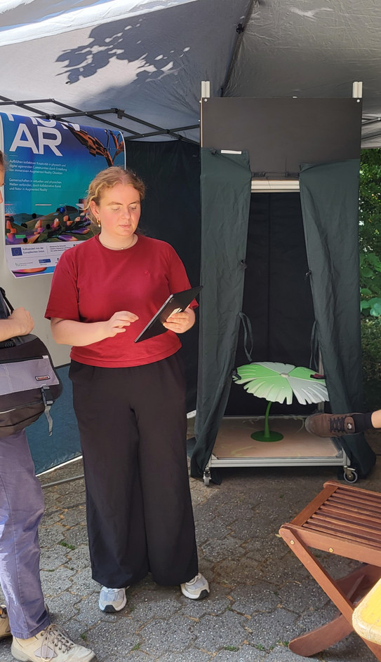 Eine junge Frau im roten T-Shirt steht an einem sonnigen Tag unter einem Pavillon und blickt auf ein Tablet in ihren Händen. Im Hintergrund hängt ein Poster mit der Aufschrift „flow AR“. Direkt daneben befindet sich eine dunkle, geöffnete Kabine. Darin liegt ein beleuchtetes, flaches Objekt in Form einer Blume auf dem Boden.