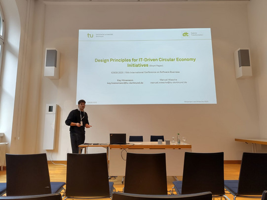 The image captures a presentation taking place in a bright conference room with high ceilings and wooden flooring. In the center, a large projection screen displays the title slide of a presentation. The slide features the logo of "tu technische universität dortmund" in the top left corner and the "dt Digitale Transformation" logo in the top right. The title reads "Design Principles for IT-Driven Circular Economy Initiatives (Short Paper)" in green text. Below the title, smaller grey text indicates "ICSOB 2025 | 16th International Conference on Software Business." The authors, Kay Hönemann and Manuel Wiesche, are listed at the bottom with their TU Dortmund email addresses.  To the left of the screen stands a male speaker with dark hair and glasses, dressed in a black sweater and dark trousers. He wears a white lanyard around his neck, holds a presenter remote, and is gesturing while looking towards the audience. In front of the screen is a light wooden table holding an open laptop, various cables, two small speakers, water bottles, and glasses. On the left wall, there are large windows and white wall-mounted speakers.