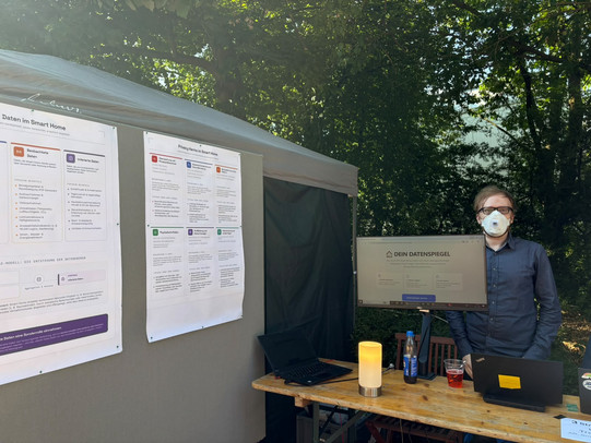 Ein junger Mann mit Brille und FFP2-Maske steht im Freien hinter einem Holztisch. Auf dem Tisch befinden sich ein Monitor mit der Webseite „DEIN DATENSPIEGEL“, zwei aufgeklappte Laptops, Getränke und eine orange leuchtende Zylinderlampe. Links neben dem Stand steht eine graue Stellwand mit zwei großen Postern zum Thema Datenschutz im Smart Home.