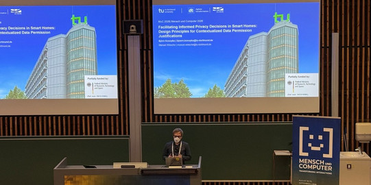 A speaker wearing glasses, an FFP2 mask, and dark clothing presents at a lectern at the Mensch und Computer 2026 conference. The projection screens behind him show the title slide for his paper on design principles for privacy decisions in smart homes. The slide displays the TU Dortmund University logo, author names Björn Konopka and Manuel Wiesche, and a photograph of the university's main high-rise building against a blue sky.