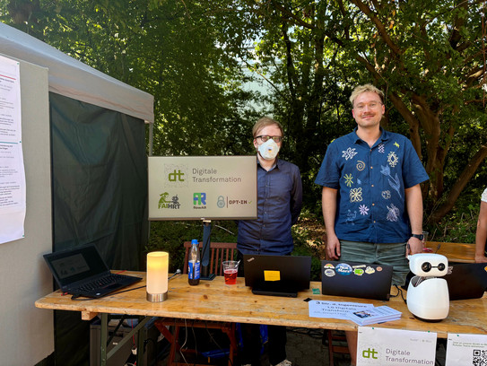 Zwei Männer stehen im Freien hinter einem Informationsstand aus Holz. Der linke Mann trägt eine FFP2-Maske, der rechte Mann ein dunkelblaues Hemd mit hellen Mustern. In der Mitte des Tisches zeigt ein Monitor das Logo „dt Digitale Transformation“ mit drei kleineren Projektlogos. Auf dem Tisch stehen zusätzlich zwei Laptops, eine orangefarbene Lampe, Informationsflyer und ein kleiner weißer humanoider Roboter.
