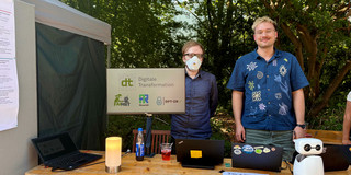 Two men stand outdoors behind a wooden information booth. The man on the left wears an FFP2 mask; the man on the right wears a dark blue shirt with light patterns. A monitor in the center of the table displays the "dt Digitale Transformation" logo and three smaller project logos. The table also holds two laptops, an orange lamp, informational flyers, and a small white humanoid robot.