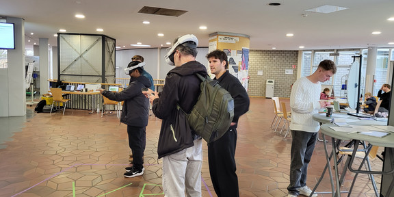 The horizontal photograph depicts a scene in a spacious, well-lit foyer of a public building, likely a university or school due to the architecture. The floor features distinctive reddish-brown hexagonal tiling throughout. In the center foreground, two individuals are actively using white Virtual Reality headsets and holding controllers. They are standing inside a designated square area marked on the floor with purple tape. Inside this square, strips of green tape spell out the large letters "VR". The person closest to the viewer is wearing a dark jacket, light grey jeans, and carrying an olive-green backpack. To their left, the second VR user is dressed in dark clothing. A third man, dressed entirely in black, stands just outside the designated zone, watching the users. To the right of the image, a young man with light hair, wearing a white cable-knit sweater and dark grey jeans, leans against a high round table. He is looking down at papers or a device, with a coffee cup sitting on the table in front of him. The background reveals a glass-walled structure on the left, tables set up with laptops and yellow chairs, and a central circular pillar covered in flyers and notices. Further back, exposed brick walls, large windows letting in daylight, and other people sitting at tables are visible. The ceiling is white with recessed circular lights.