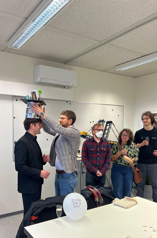 The photo captures a celebration in a brightly lit office or seminar room, featuring a group of five people, likely marking an academic milestone like a PhD defense. On the left side, the main focus is on a smiling man dressed in a black suit and black shirt. He is standing still while another man places a custom-made graduation cap (mortarboard) on his head. The man placing the hat wears a grey plaid blazer, blue jeans, and glasses. The hat itself is heavily decorated; the cylindrical part is covered in small printed photos, and the top features miniature objects, including a small palm tree and a frame. To the right of this pair, three onlookers observe the scene. Closest to the center is a man wearing a red plaid shirt, jeans, and glasses, who is also wearing a white face mask and has his hands clasped. Next to him stands a woman with curly hair and glasses, wearing a yellow and green patterned sweater; she holds a glass of sparkling wine and a brown bag. On the far right stands a tall man in a black t-shirt and grey trousers, also holding a glass of sparkling wine and smiling. In the immediate foreground, resting on a white table, is a white balloon with the text "Congrats Class of 2025" printed in a festive font. The background features a whiteboard, a wall-mounted air conditioning unit, and a display area featuring a large model of a construction crane or excavator (likely a Lego Technic model). The room has a standard drop ceiling with fluorescent lighting.