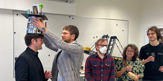 The photo captures a celebration in a brightly lit office or seminar room, featuring a group of five people, likely marking an academic milestone like a PhD defense. On the left side, the main focus is on a smiling man dressed in a black suit and black shirt. He is standing still while another man places a custom-made graduation cap (mortarboard) on his head. The man placing the hat wears a grey plaid blazer, blue jeans, and glasses. The hat itself is heavily decorated; the cylindrical part is covered in small printed photos, and the top features miniature objects, including a small palm tree and a frame.  To the right of this pair, three onlookers observe the scene. Closest to the center is a man wearing a red plaid shirt, jeans, and glasses, who is also wearing a white face mask and has his hands clasped. Next to him stands a woman with curly hair and glasses, wearing a yellow and green patterned sweater; she holds a glass of sparkling wine and a brown bag. On the far right stands a tall man in a black t-shirt and grey trousers, also holding a glass of sparkling wine and smiling.  In the immediate foreground, resting on a white table, is a white balloon with the text "Congrats Class of 2025" printed in a festive font. The background features a whiteboard, a wall-mounted air conditioning unit, and a display area featuring a large model of a construction crane or excavator (likely a Lego Technic model). The room has a standard drop ceiling with fluorescent lighting.