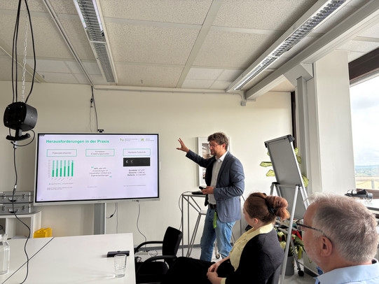The photo captures a presentation or workshop scenario inside a brightly lit seminar room. The central figure is a male presenter standing to the right of a large flat-screen monitor mounted on a stand. He is wearing glasses, a blue blazer over a light-colored shirt, and jeans. He gestures with his right hand toward the screen while maintaining eye contact with the audience. The screen displays a presentation slide with the green headline "Herausforderungen in der Praxis" (Challenges in Practice). The slide features logos for TU Dortmund and "dt," a green bar chart on the left, a quote in the center, and a technical image (likely Augmented Reality hardware) on the right with specifications such as field of view and weight.  In the right foreground, two audience members are seated in profile, attentively watching the presentation: on the far right, a man with short gray hair, glasses, and a light blue shirt, and next to him, a woman with reddish-brown hair styled in a bun, wearing a black top with a yellow collar or scarf. The room has a functional design with a suspended grid ceiling containing long fluorescent lights. On the left side of the image, a black power outlet cube hangs from the ceiling on a chain, positioned above a white shelf unit holding several silver metal cases. To the right, behind the presenter, a flip chart and a potted plant stand in front of large windows that offer a view of a cloudy sky and a cityscape. Water glasses and a smartphone are placed on the white table in the immediate foreground.