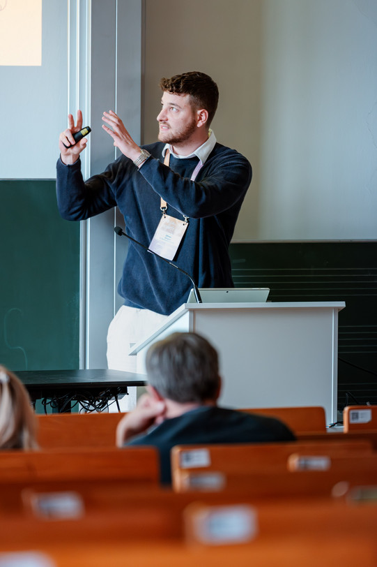 Eine Fotografie aus der Perspektive des Publikums in einem hellen Hörsaal. Im Zentrum steht ein Mann mit dunklem, lockigem Haar und Bart an einem weißen Rednerpult. Er trägt einen dunkelblauen, gerippten Pullover über einem weißen Hemd, eine helle Hose und ein Umhängeband mit Konferenz-Namensschild. Er blickt nach links über die Zuhörer hinweg und gestikuliert lebhaft mit beiden erhobenen Händen, wobei er in der rechten Hand einen schwarzen Präsentationsklicker hält. Auf dem schlichten Pult stehen ein Laptop und ein Schwanenhalsmikrofon. Im unscharfen Vordergrund sind die Hinterköpfe von zwei zuhörenden Personen sowie Reihen von braunen Holzbanken zu sehen. Im Hintergrund befinden sich eine grüne Kreidetafel und eine stark beleuchtete, weiße Projektionsfläche.