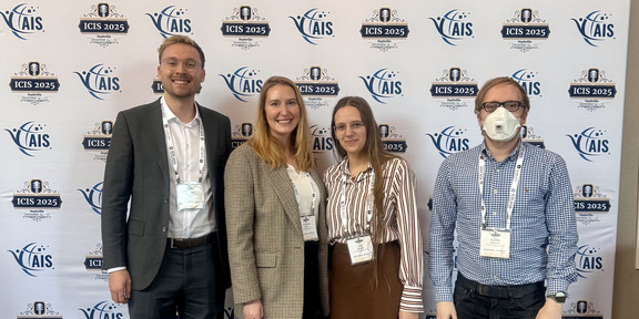 ICIS 2025 Group photo The image is a full-body group photo showing four people standing side-by-side facing the camera in front of a conference backdrop. The backdrop is white and features a banner at the top reading "ICIS 2025" in large dark blue letters, followed by "Nashville Tennessee December 14 – 17". The banner includes graphics of a guitar headstock on the left and an orange guitar neck on the right containing the hashtag #ICIS2025. Below the banner, the wall features a repeating step-and-repeat pattern of logos for "AIS" (Association for Information Systems) and "ICIS 2025". From left to right: The first person is a man with short blond hair and glasses, wearing a dark grey suit jacket, a white button-down shirt, dark trousers, and brown leather dress shoes. Next to him is a woman with long blond hair wearing a beige checked blazer, a white top, black trousers, and light-colored sneakers. Beside her stands a woman with long brown hair and glasses, wearing a vertically striped shirt in white and reddish-brown, a brown midi skirt, dark tights, and tan boots with fur trim. On the far right is a man with short hair and glasses wearing a white face mask. He is dressed in a blue checkered button-down shirt, dark jeans, and brown boots. All four individuals are wearing conference name badges on lanyards around their necks. They are standing on a carpet patterned with large, abstract leaf shapes in shades of blue and green.