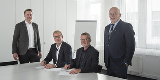 Dr. Ronald Kriedel and Prof. Andreas Liening are standing at a table. Albrecht Ehlers and Guido Baranowski sit at the table and sign contracts