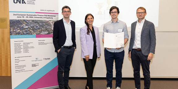 vier Personen (drei Männer und eine Frau) stehen vor einem Aufsteller des Workshops 2nd European Sustainable Finance PhD