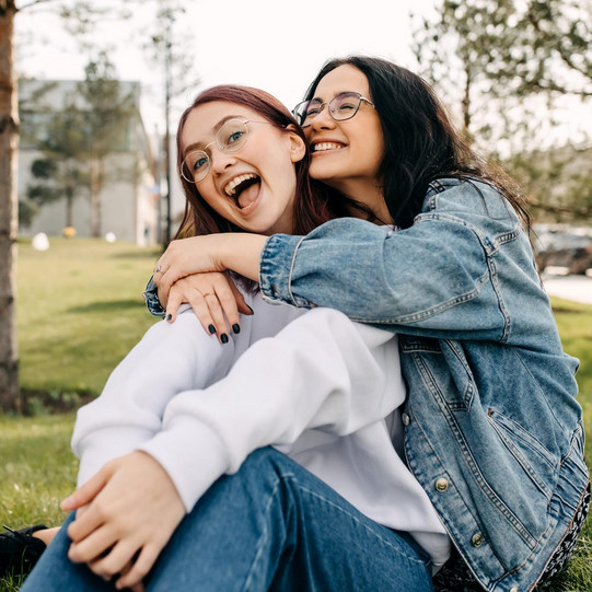 Two young women sitting in the grass, laughing and hugging each other.