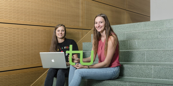 Two young women are sitting on a staircase. One is holding a laptop, and the other is holding the TU Dortmund logo.