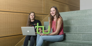 Two young women are sitting on a staircase. One is holding a laptop, and the other is holding the TU Dortmund logo. 