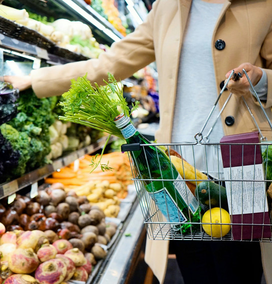 Frau in heller Kleidung hält einen Einkaufskofb mit einer Flasche und Obst und Gemüse. Sie steht vor einem Gemüseregal im Supermarkt. 