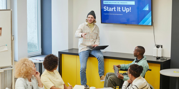 A man is giving a presentation to a group at a conference table; the words 'Start it up!' appear on a screen behind him.