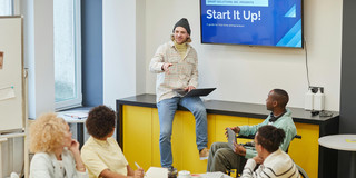 A man is giving a presentation to a group at a conference table; the words 'Start it up!' appear on a screen behind him.