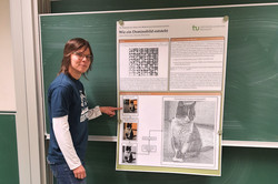 Woman standing in front of a blackboard with a poster hanging on it. The poster explains how domino pictures are created.