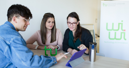 Zwei Studentinnen und ein Student sitzen zusammen und schauen auf ein Tablet. 