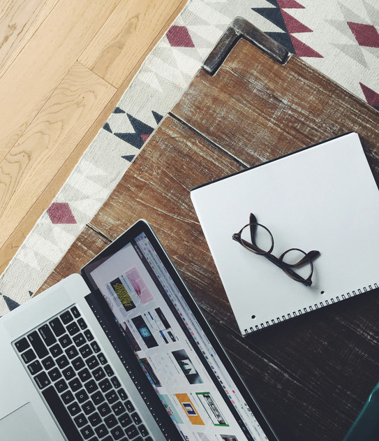 Laptop on wooden table, small square pictures on screen. Behind it a notebook with glasses.
