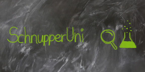 The text "SchnupperUni" written in green chalk on a not quite cleanly wiped blackboard, next to it a painted magnifying glass and a half-filled test tube
