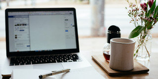 Foto: geöffnetes Notizbuch vor Laptop, daneben Kaffeetasse und Blumen