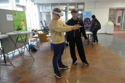 Junger Mann mit VR Headset und Controllern. Neben ihm ein zweiter junger Mann, der zusieht.