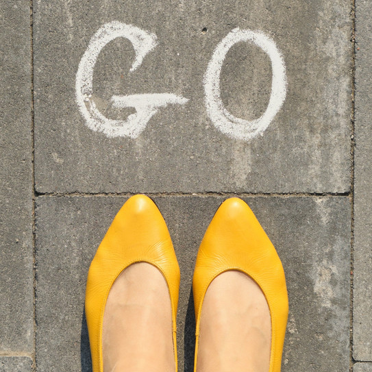 Foto: Frauenfüße in gelben Ballerinas, auf Pflastersteine, vor den Füßen steht in Kreide das Wort "Go".