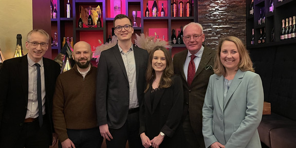 TMC Award Ceremony 2022 Photo of the award winners Silas Fischer and Celine Daute, the donors Christian Lützenrath and Steffen Leininger as well as the supervising university lecturers Andreas Hoffjan and Christiane Pott in front of colorfully illuminated, modern shelves in which selected wines are presented.