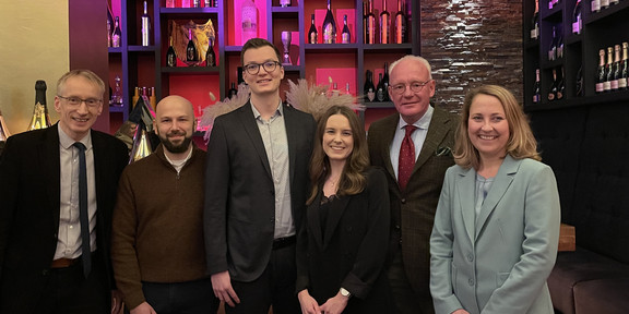 TMC Preisverleihung 2022 Foto der Preisträger Silas Fischer und Celine Daute, die Stifter Christian Lützenrath und Steffen Leininger sowie die betreuenden Hochschullehrer Andreas Hoffjan und Christiane Pott vor bunt angestrahlten, modernen Regalen, in denen ausgesuchte Weine präsentiert werden.