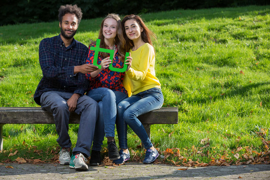 Three students sitting on a bench and holding a green TU logo in their hands together.