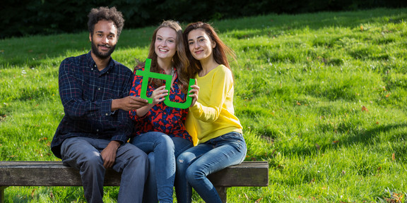 Three students sitting on a bench and holding a green TU logo in their hands together.
