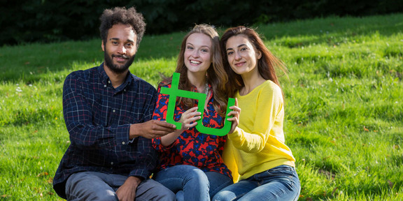 Drei Studierende sitzen auf einer Bank und halten gemeinsam ein grünes TU-Logo in den Händen.