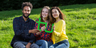 Drei Studierende sitzen auf einer Bank und halten gemeinsam ein grünes TU-Logo in den Händen.