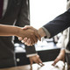 Two people shake hands. The handshake is shown in detail, in the background are two people in suits.