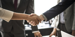 Two people shake hands. The handshake is shown in detail, in the background are two people in suits.