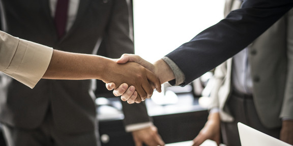 Two people shake hands. The handshake is shown in detail, in the background are two people in suits.