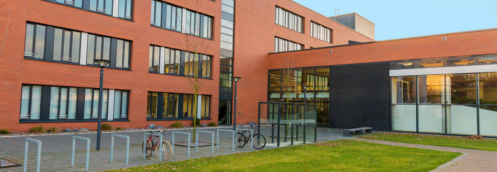 Seminarraumgebäude: Ein rotes dreistöckiges Gebäude mit schwarz gerahmten Fenstern, von der Seite fotografiert. Davor eine Wiese und Fahrradständer sowie Straßenlaternen.