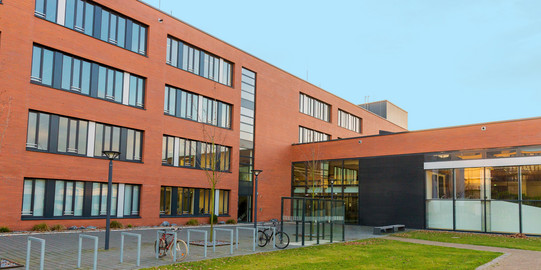 Seminarraumgebäude: Ein rotes dreistöckiges Gebäude mit schwarz gerahmten Fenstern, von der Seite fotografiert. Davor eine Wiese und Fahrradständer sowie Straßenlaternen.