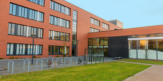 Seminarraumgebäude: Ein rotes dreistöckiges Gebäude mit schwarz gerahmten Fenstern, von der Seite fotografiert. Davor eine Wiese und Fahrradständer sowie Straßenlaternen.